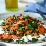 A rustic ceramic plate holds a vibrant lentil and roasted carrot salad. The dish features tender, caramelized carrot slices mixed with black lentils and finely chopped green onions. Fresh herbs are sprinkled on top, adding a burst of color. In the softly blurred background, a bottle of olive oil, a bowl, and a folded blue napkin rest on a textured surface.